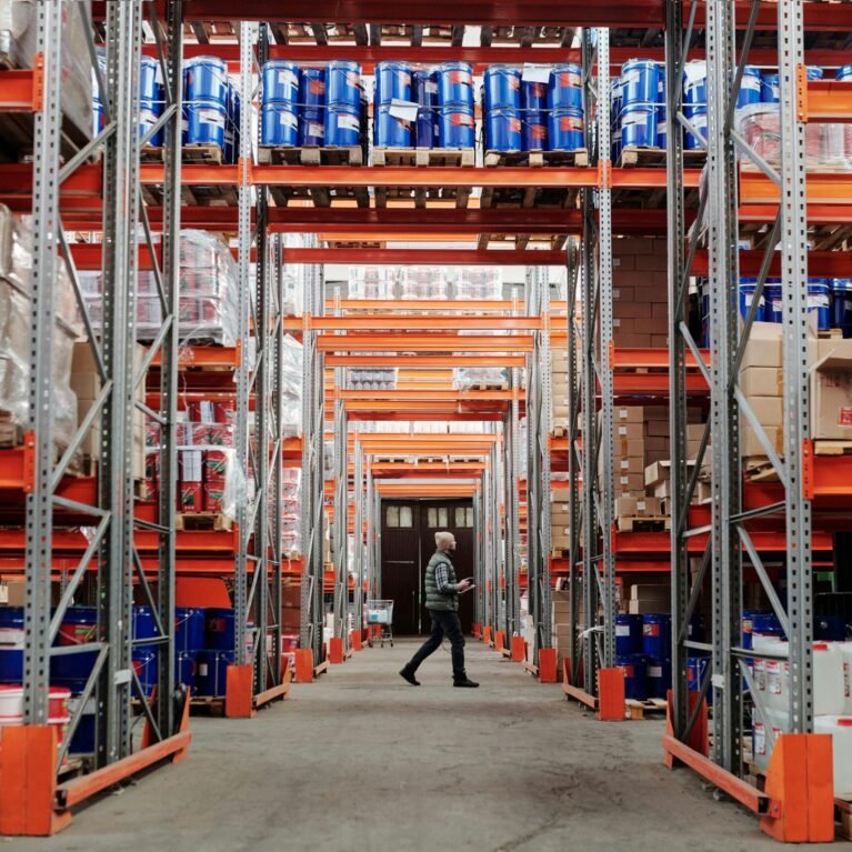 A man walking through a large industrial warehouse with stacked shelves filled with goods and products.